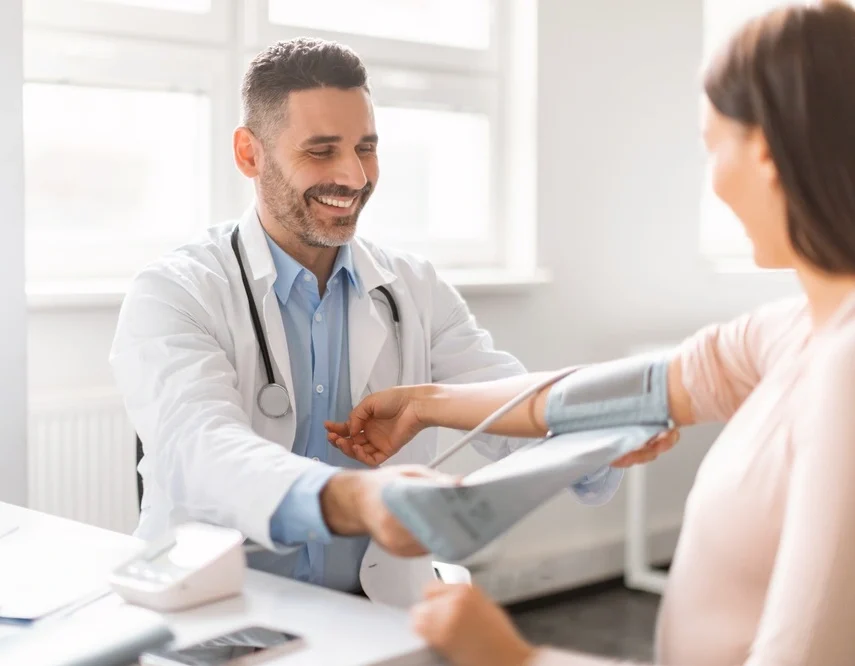 Male doctor measuring arterial blood pressure of young pregnant lady using cuff, sitting in clinic office