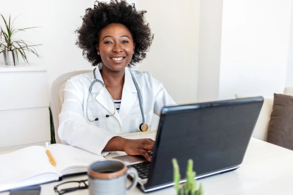 Portrait of smiling female doctor wearing white coat with stethoscope sitting behind desk in office.